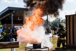 Photo of a Little York FD Firefighter in full bunker gear extinguishing a fire with a fire extinguisher in front of the community during a live fire demo at the 2024 Little York FD Fire Fest. Flames can be scene taller than the firefighter.